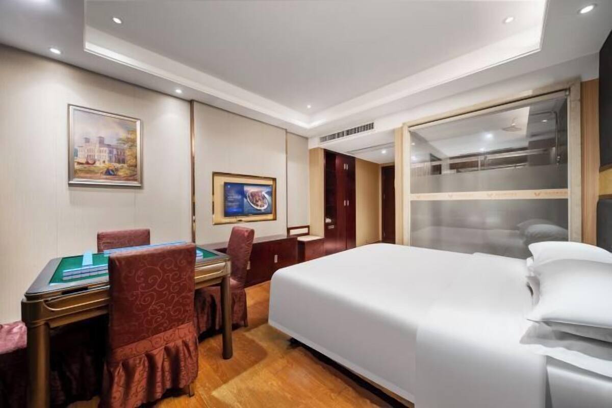 A hotel room with a bed, a desk with a green felt table, and a chair. There is a painting on the wall and a door with a glass panel.