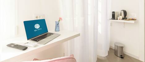 A modern room with a laptop on a desk, a remote control, a vase with a flower, a trash bin, and a shelf with a coffee maker and cups.
