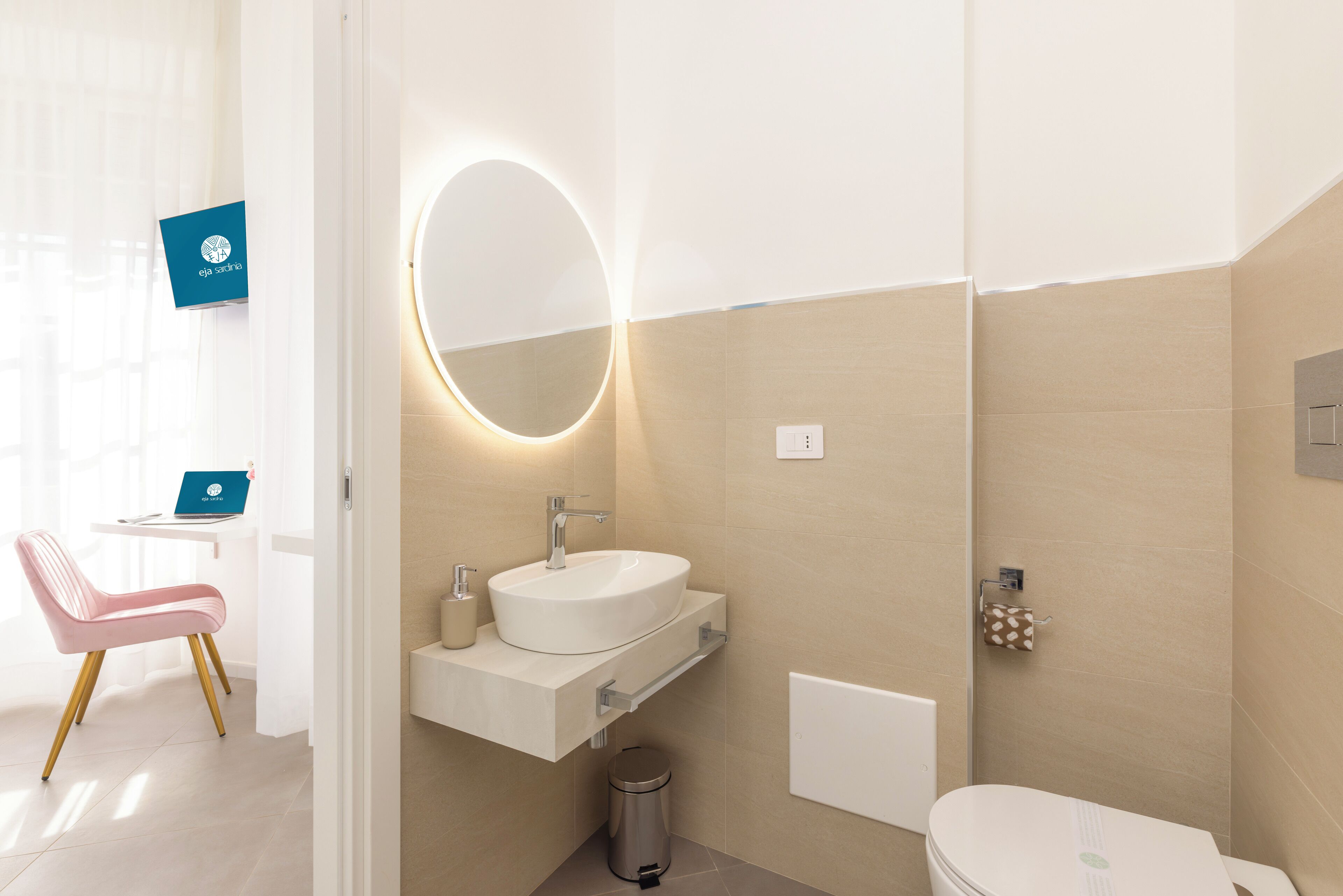 Un baño moderno con lavamanos, espejo y retrete blancos. Incluye un cesto de basura y un perchero para toallas.