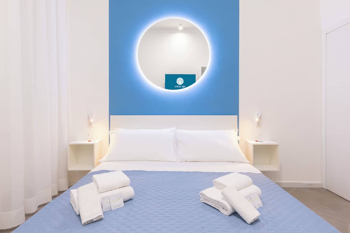 Habitación de hotel con una cama, almohadillas blancas, colcha azul acolchada, dos toallas blancas dobladas, un espejo redondo con marco azul y un pequeño letrero azul en la pared.