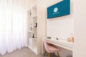 Una habitación moderna con un escritorio de cristal, una silla rosa, un estante blanco y un televisor en la pared mostrando 'eja sardinia'.