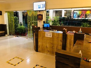 A hotel reception area with a wooden reception desk, a television screen, and a sign indicating social distancing measures.