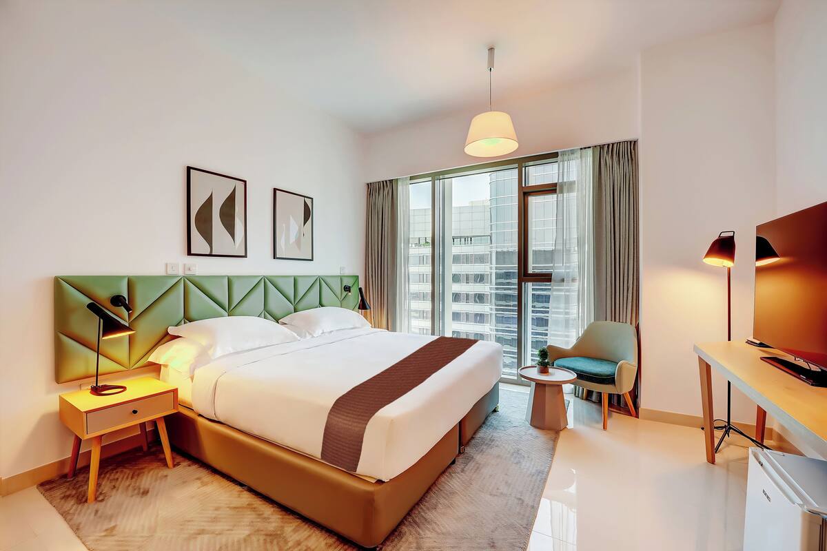 Une chambre d’hôtel moderne dotée d’un grand lit, d’un bureau, d’une chaise et offrant une vue sur la ville grâce à de grandes fenêtres.
