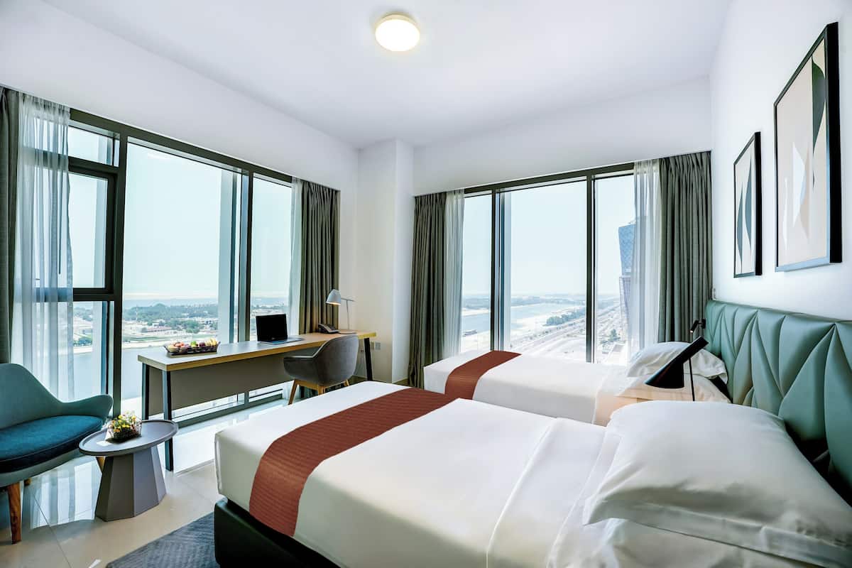 Une chambre d’hôtel avec deux lits, un bureau, une chaise et de grandes fenêtres donnant sur la ville.