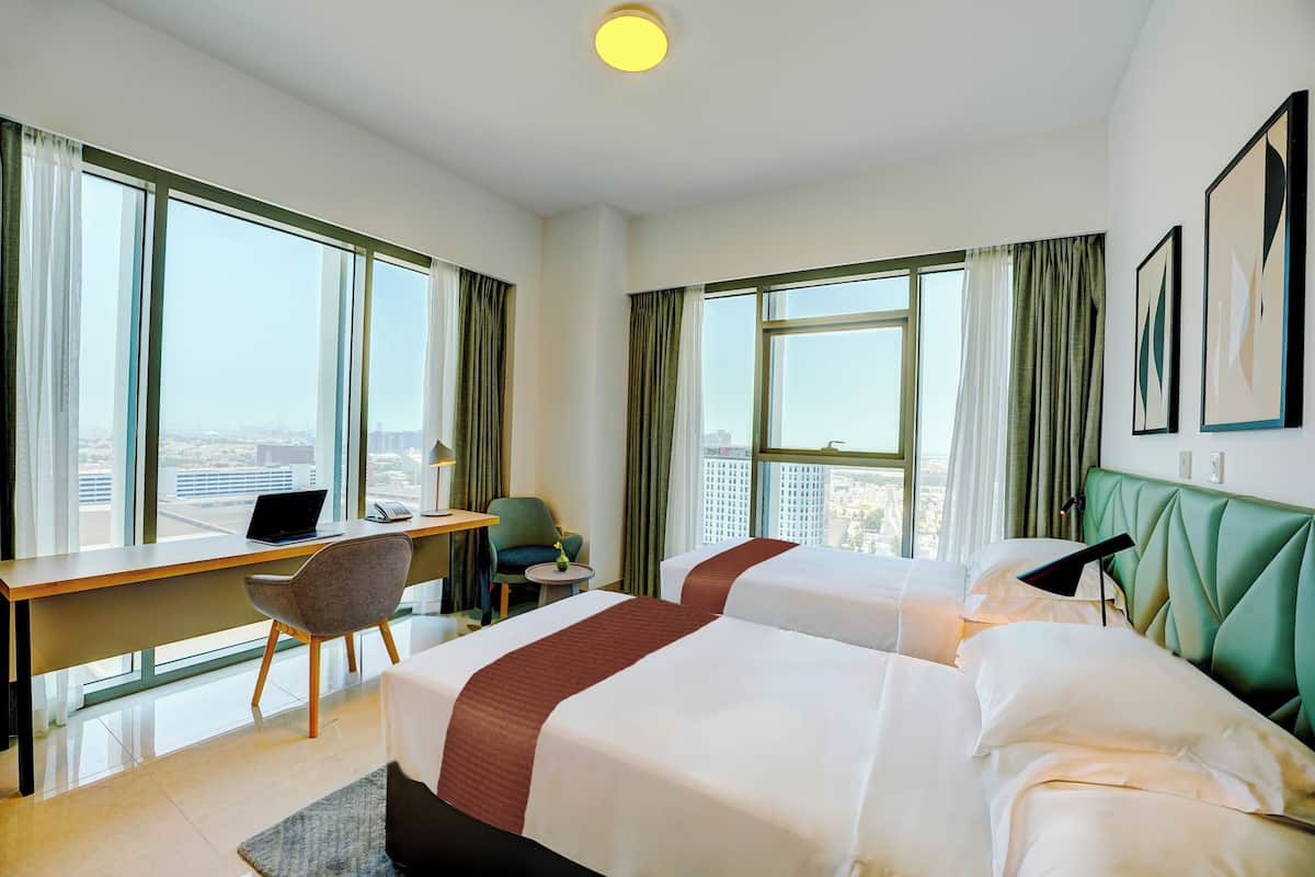 Une chambre d’hôtel avec deux lits, un bureau, une chaise et de grandes fenêtres donnant sur la ville.