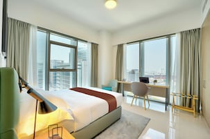 A hotel room with a large bed, a desk, and a chair, with a view of the city through large windows.