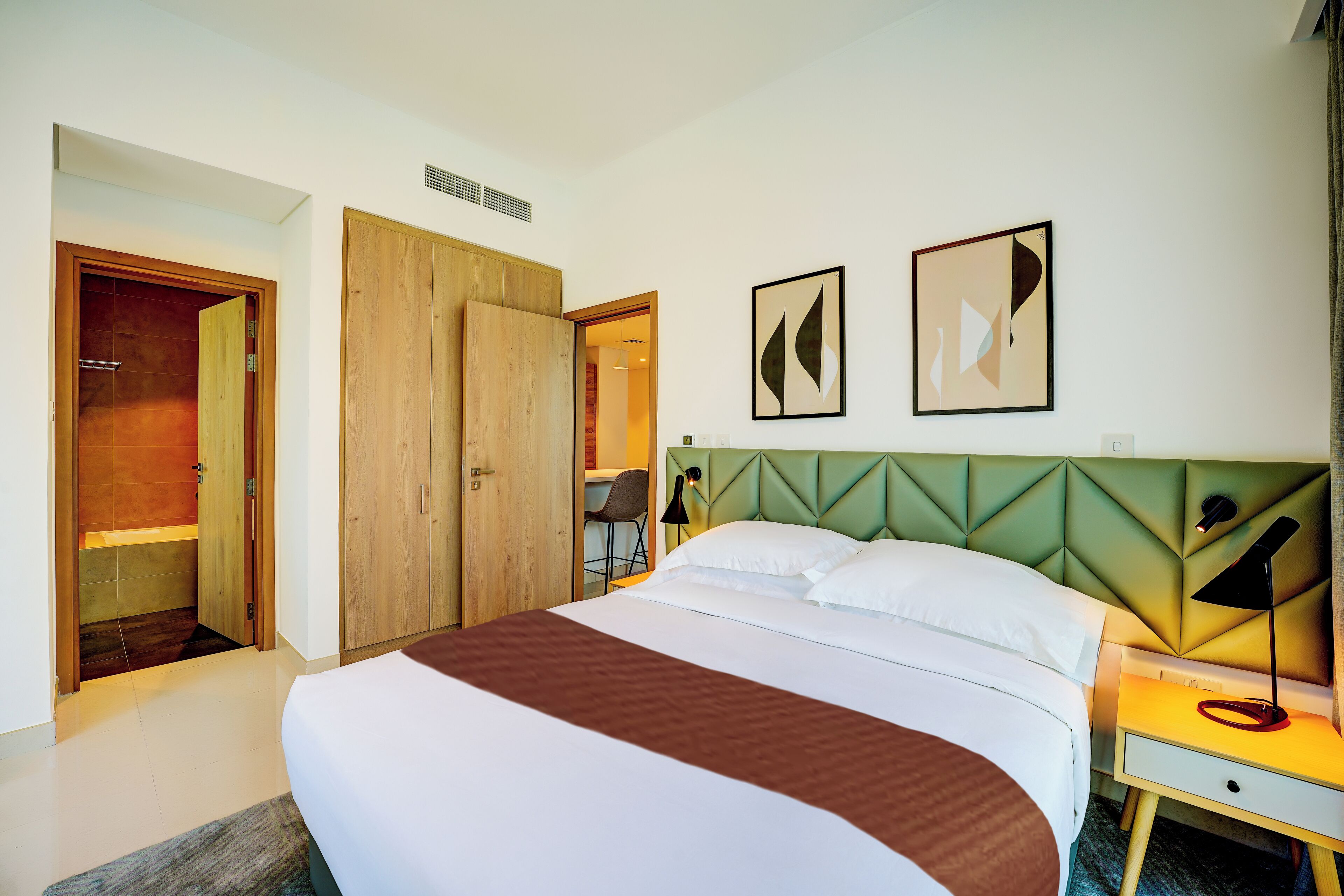 A hotel room with a large bed, a wooden wardrobe, a desk with a chair, and two abstract wall art pieces.