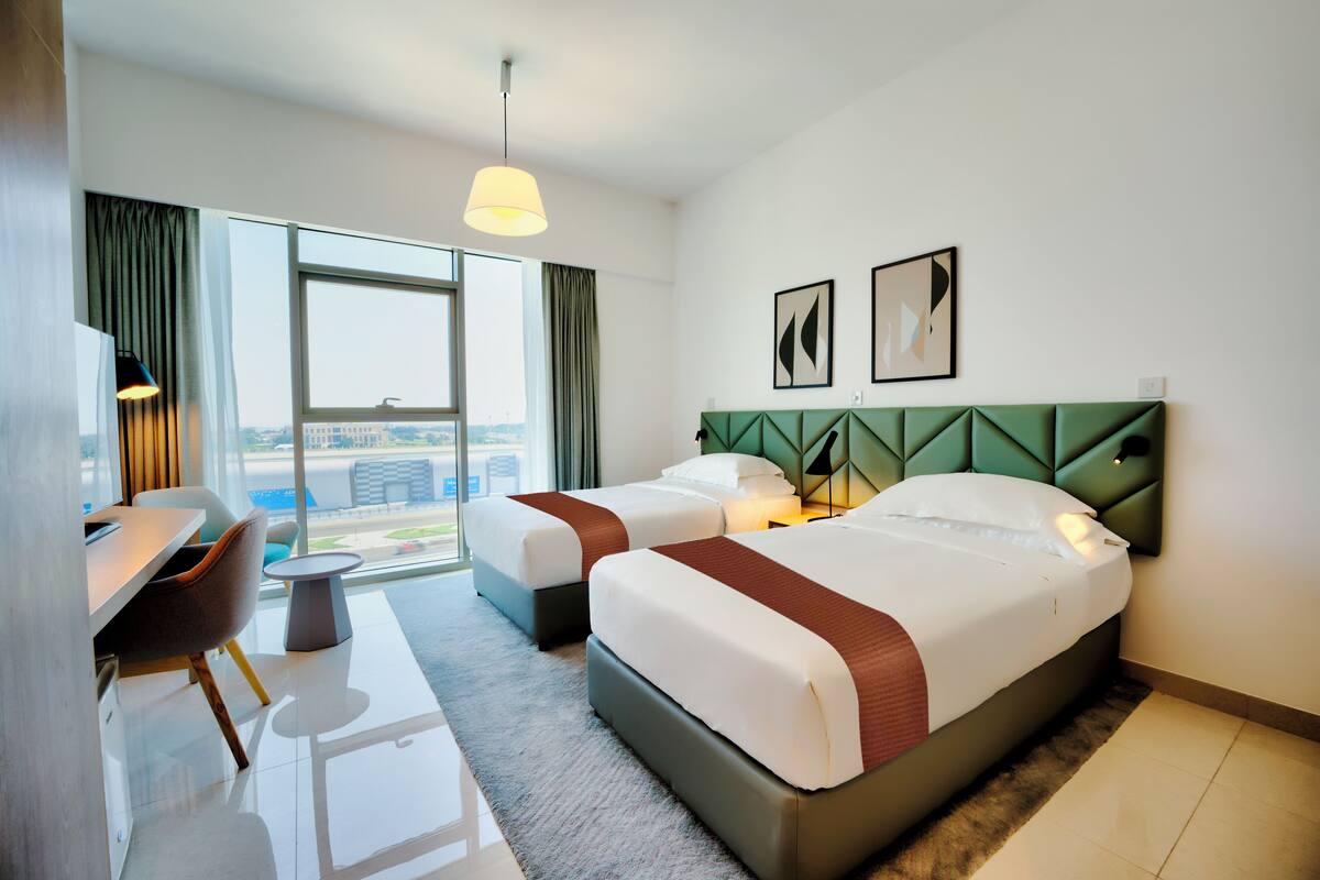 Une chambre d’hôtel avec deux lits, un bureau et une chaise. Une grande fenêtre offre une vue sur la ville.