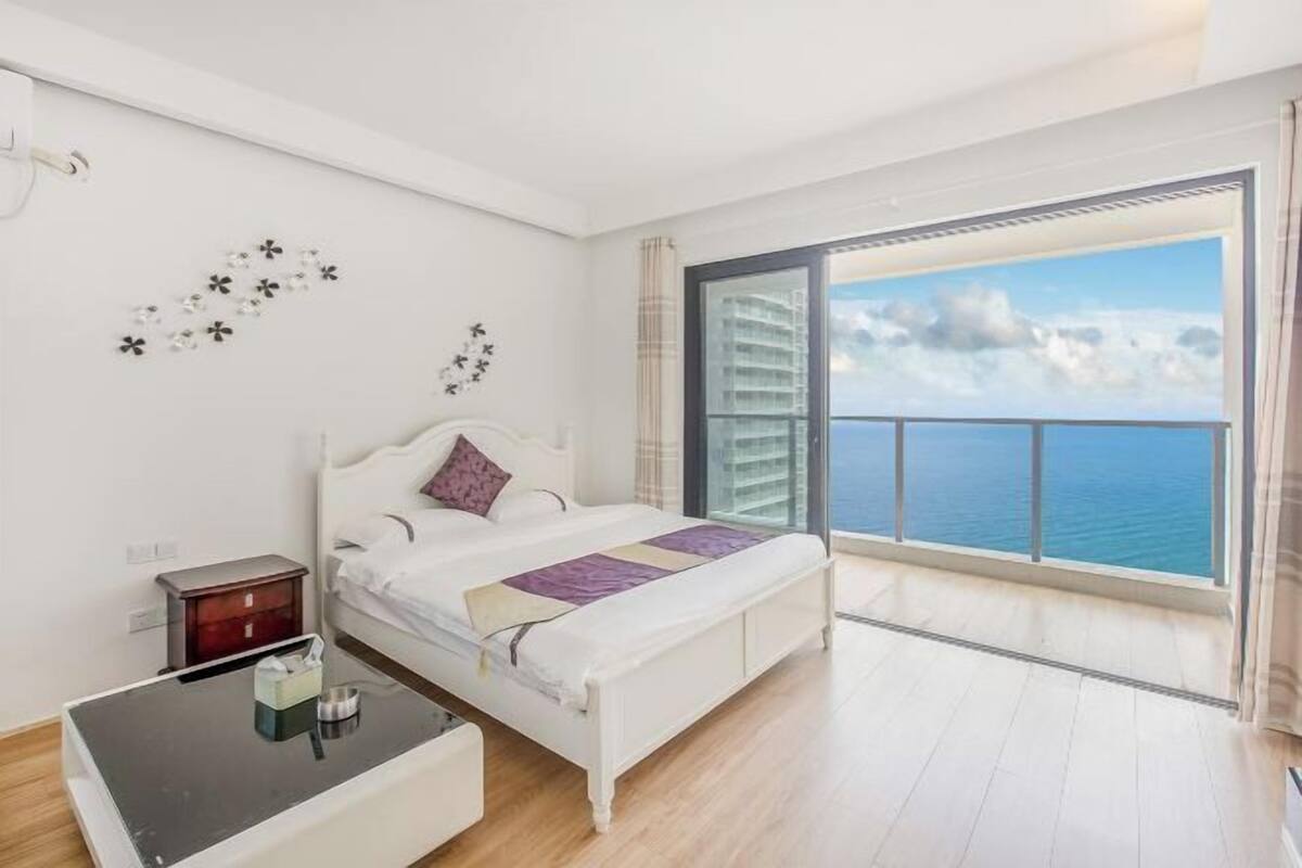 A bedroom with a bed, bedside table, and a view of the ocean through a large window.