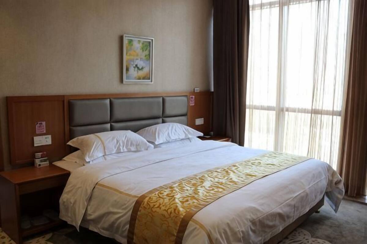 Une chambre d’hôtel avec un grand lit, une tête de lit en bois, deux tables de chevet, une petite table, une horloge murale et une fenêtre avec des rideaux.
