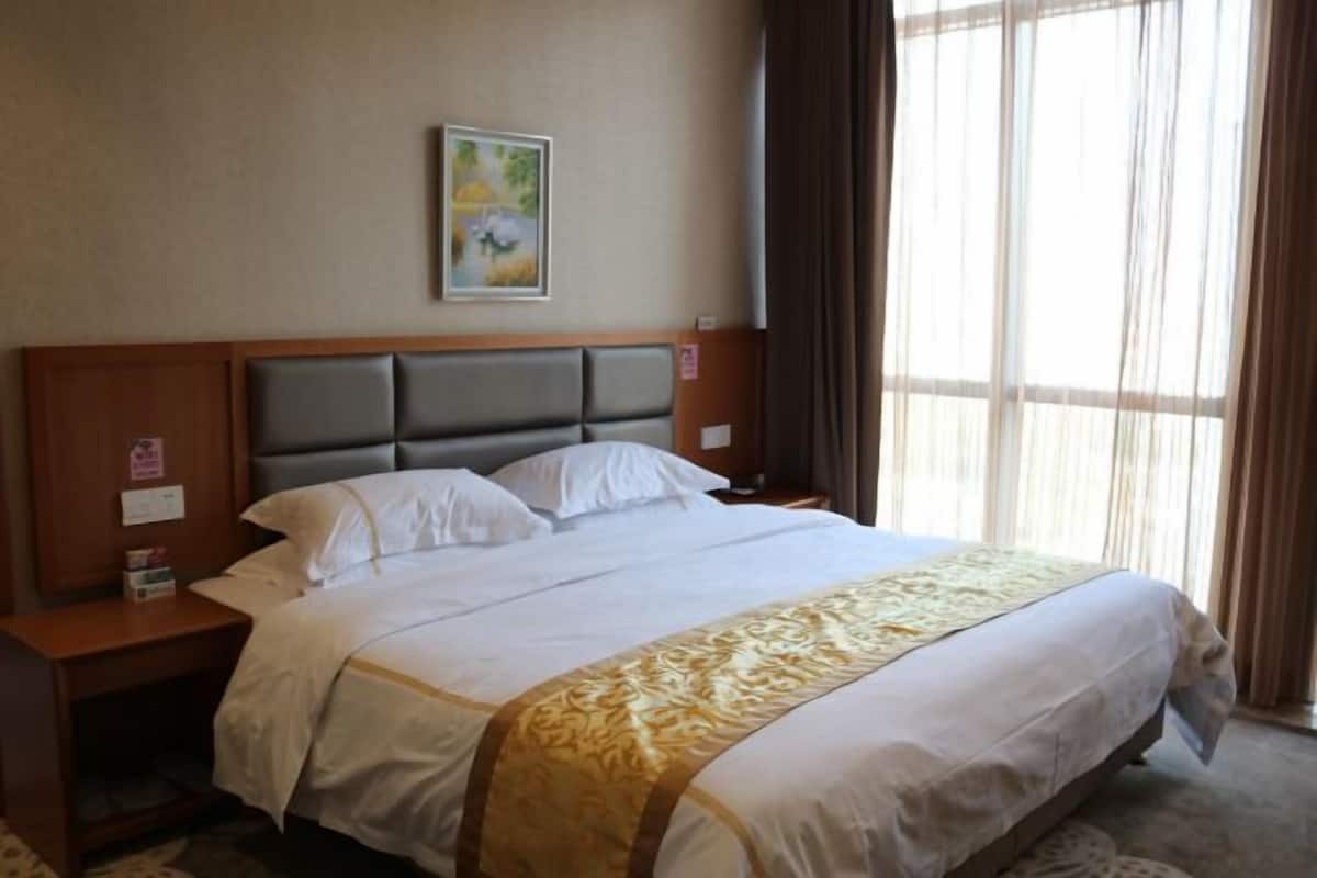 Une chambre d’hôtel avec un grand lit, une tête de lit en bois, du linge de lit blanc, une bordure décorative dorée, un tableau au mur et une fenêtre avec des rideaux.