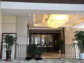 A hotel lobby with a reception desk, potted plants, and a tiled floor.