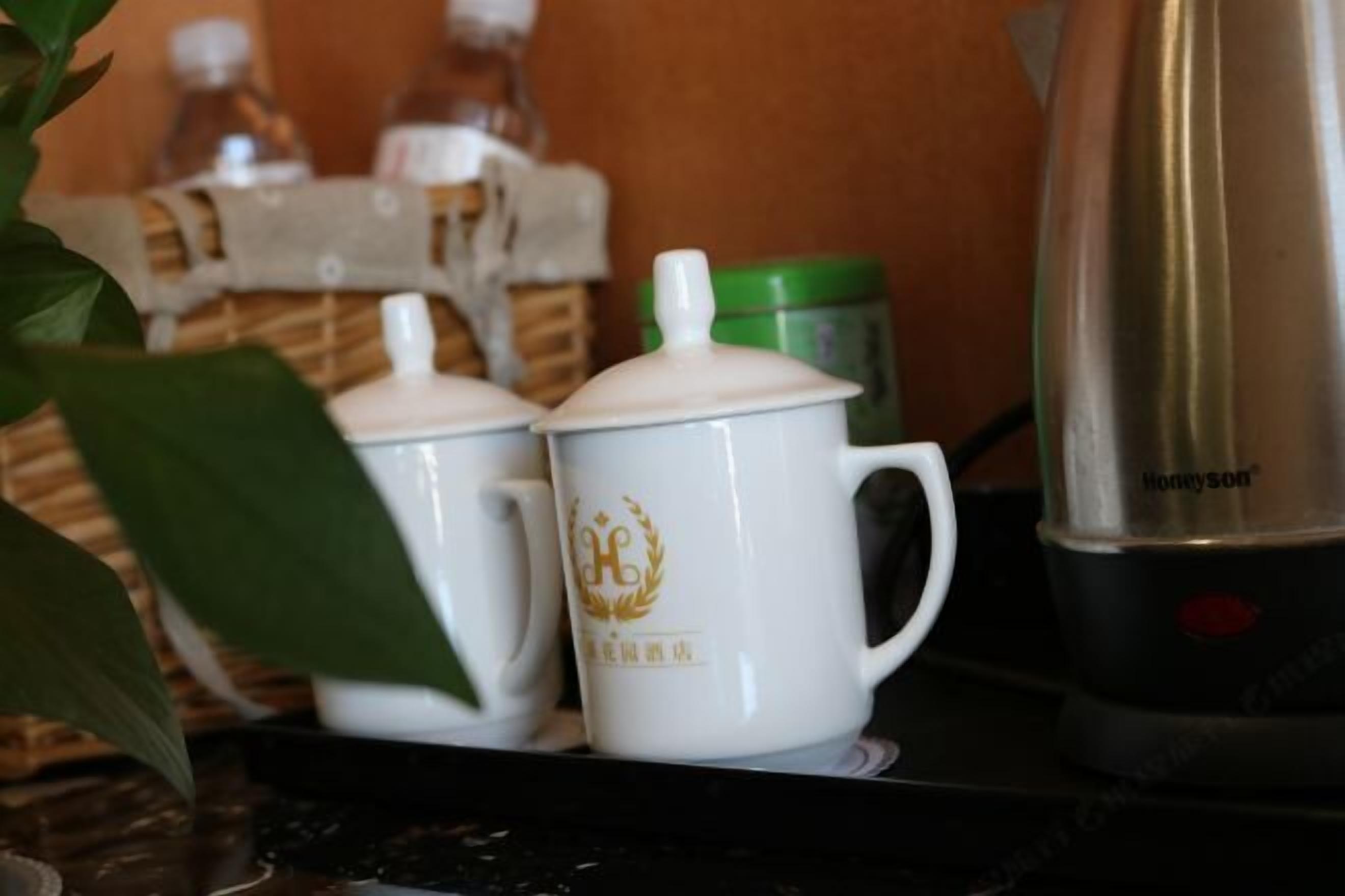 Dos tazas blancas de cerámica con emblemas dorados sobre una encimera, un recipiente verde y un hervidor eléctrico plateado.