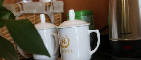 Dos tazas blancas de cerámica con emblemas dorados sobre una encimera, un recipiente verde y un hervidor eléctrico plateado.