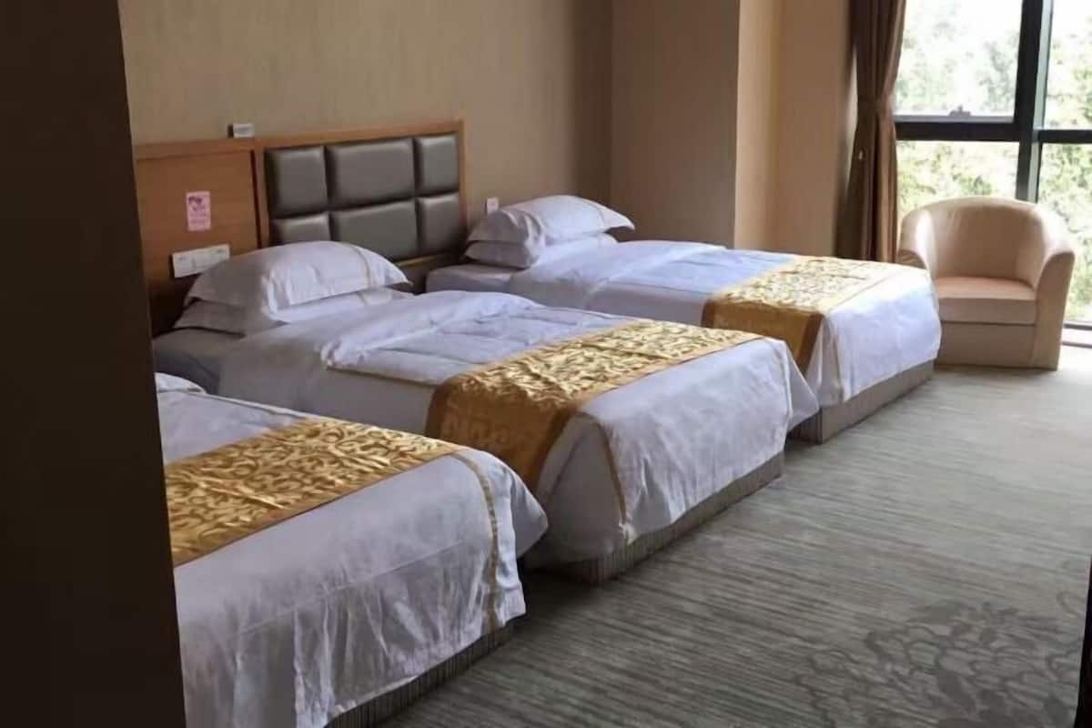 Une chambre d’hôtel avec trois lits, une tête de lit en bois, un fauteuil beige et une grande fenêtre.