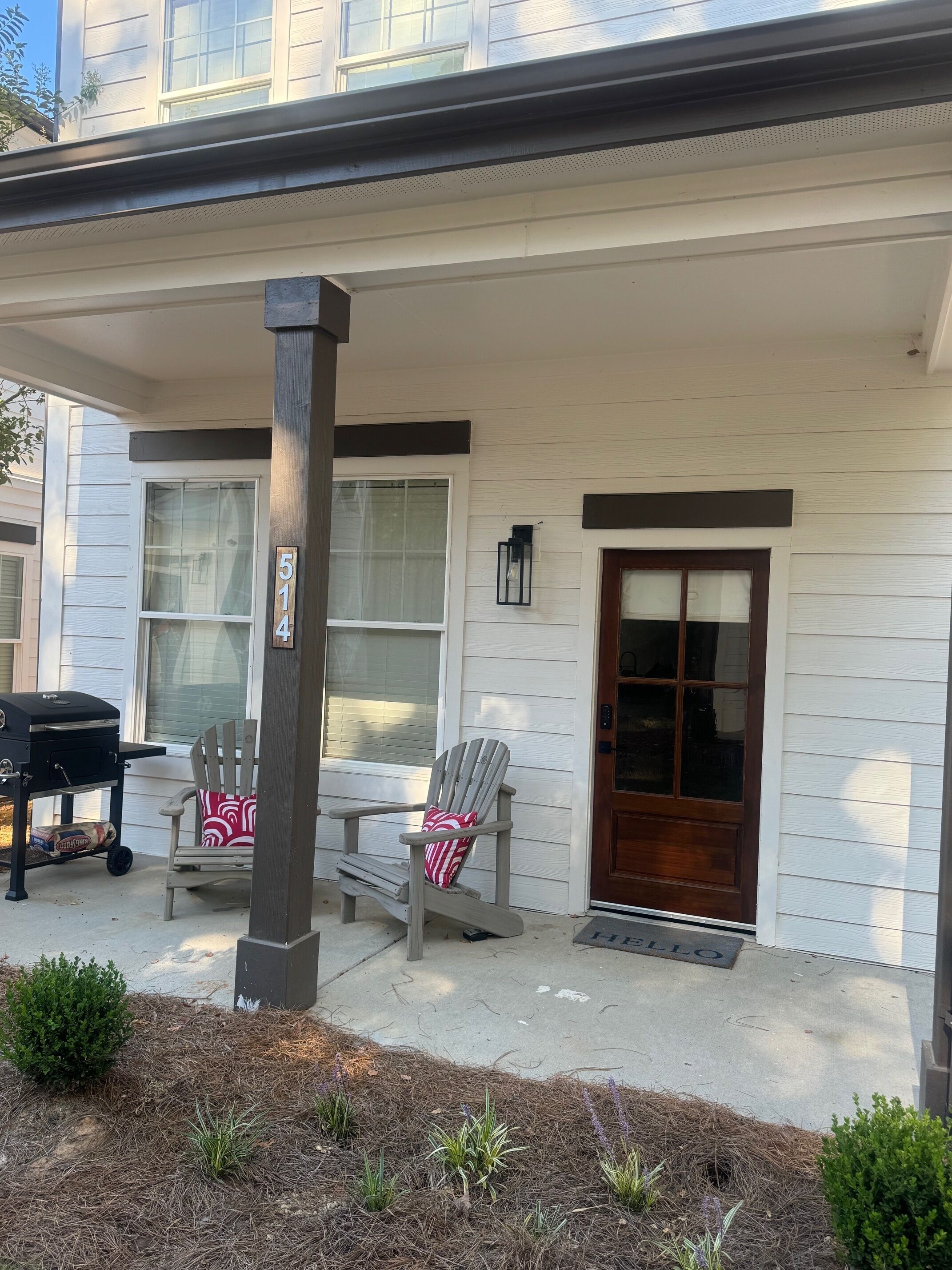 Een huis met een veranda, een barbecue, twee Adirondack-stoelen en een deur met een raam.