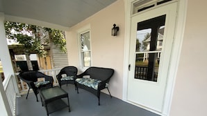 Eine überdachte Veranda mit zwei Korbstühlen und einem kleinen Tisch, einer weißen Tür und einem Fenster.