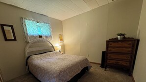 A bedroom with a bed, a dresser, a nightstand, a lamp, and a window with floral curtains. - Charming 2-bedroom house in Tiona with sunset views and everything you need. (Tiona)