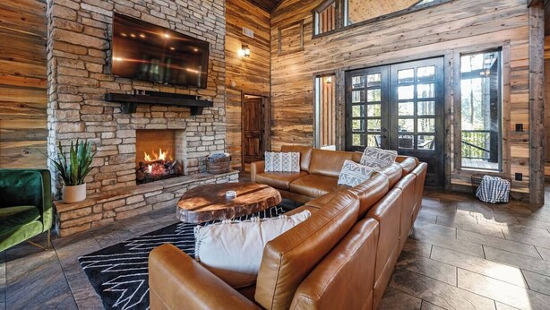 A log cabin interior with a stone fireplace, leather sofa, and large windows.