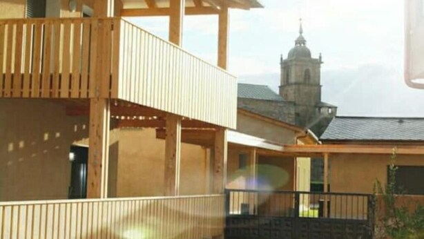 Un edificio con un balcón de madera y vistas a una torre de iglesia al fondo.