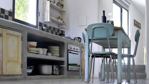 A kitchen with a dining table and chairs, a stove, and shelves with kitchenware. - Trullo Rora' - Ostuni (Puglia)