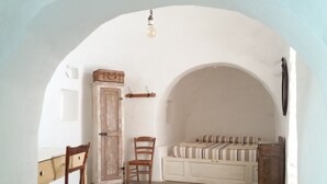 A room with a white arched doorway, a wooden chair, a bench with striped cushions, and a cabinet with a basket on top. - Trullo Rora' - Ostuni (Puglia)