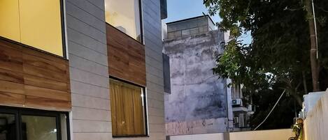 Un edificio moderno con entrada de vidrio, zona de estar al aire libre con sillas y una mesa, y un pequeño jardín con césped artificial.