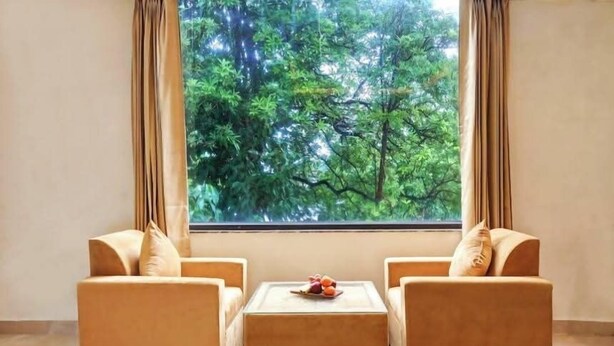 A room with two beige sofas and a square ottoman, a small table with fruit, and a large window with curtains. - Hotel Golden Era (Noida)