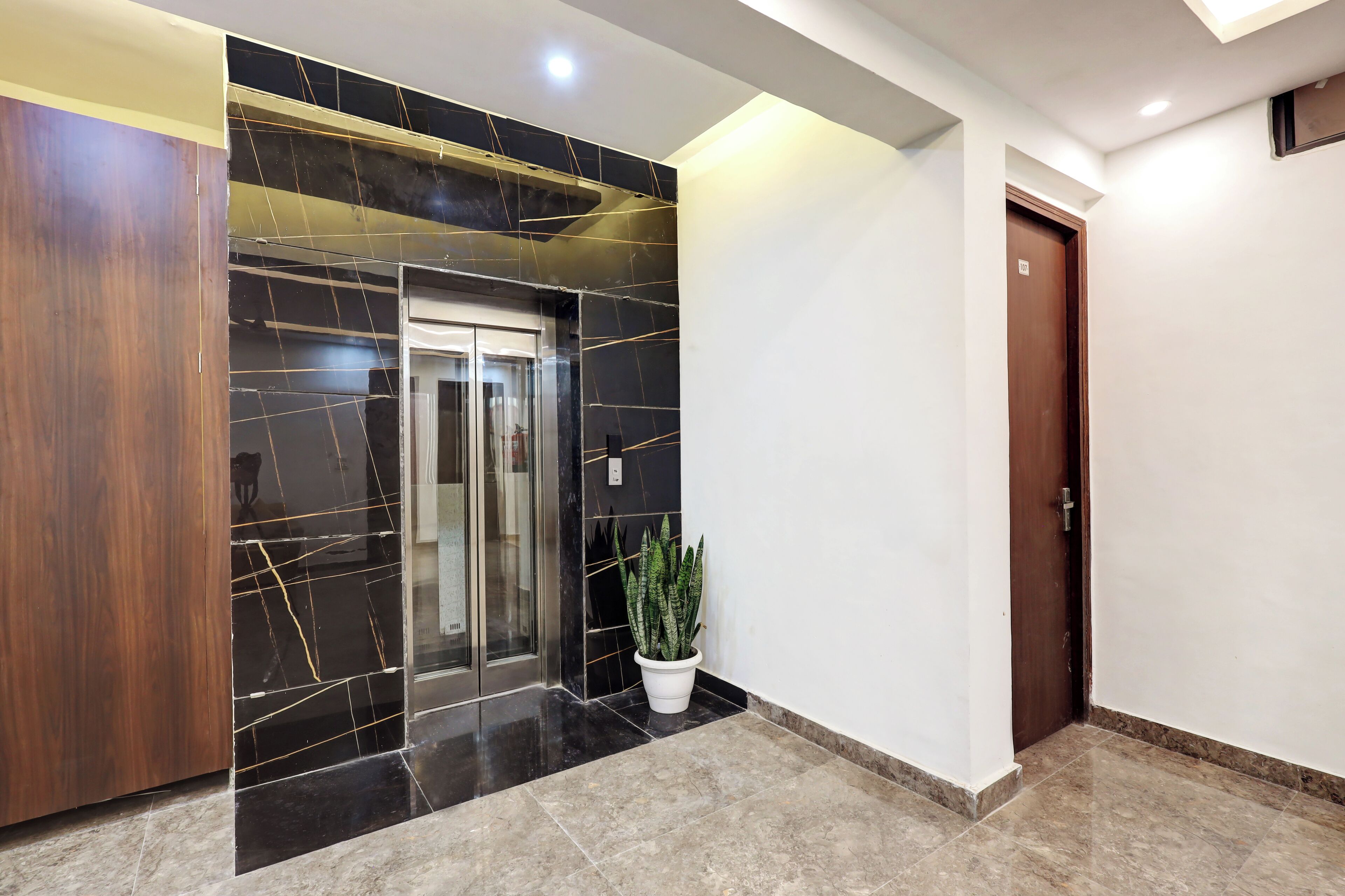 A modern interior with a wooden wardrobe, a glass elevator, and a potted plant. - Hotel Golden Era (Noida)