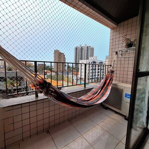 Een balkon met een hangmat, betegelde muren, uitzicht op gebouwen en een klein plankje met planten.