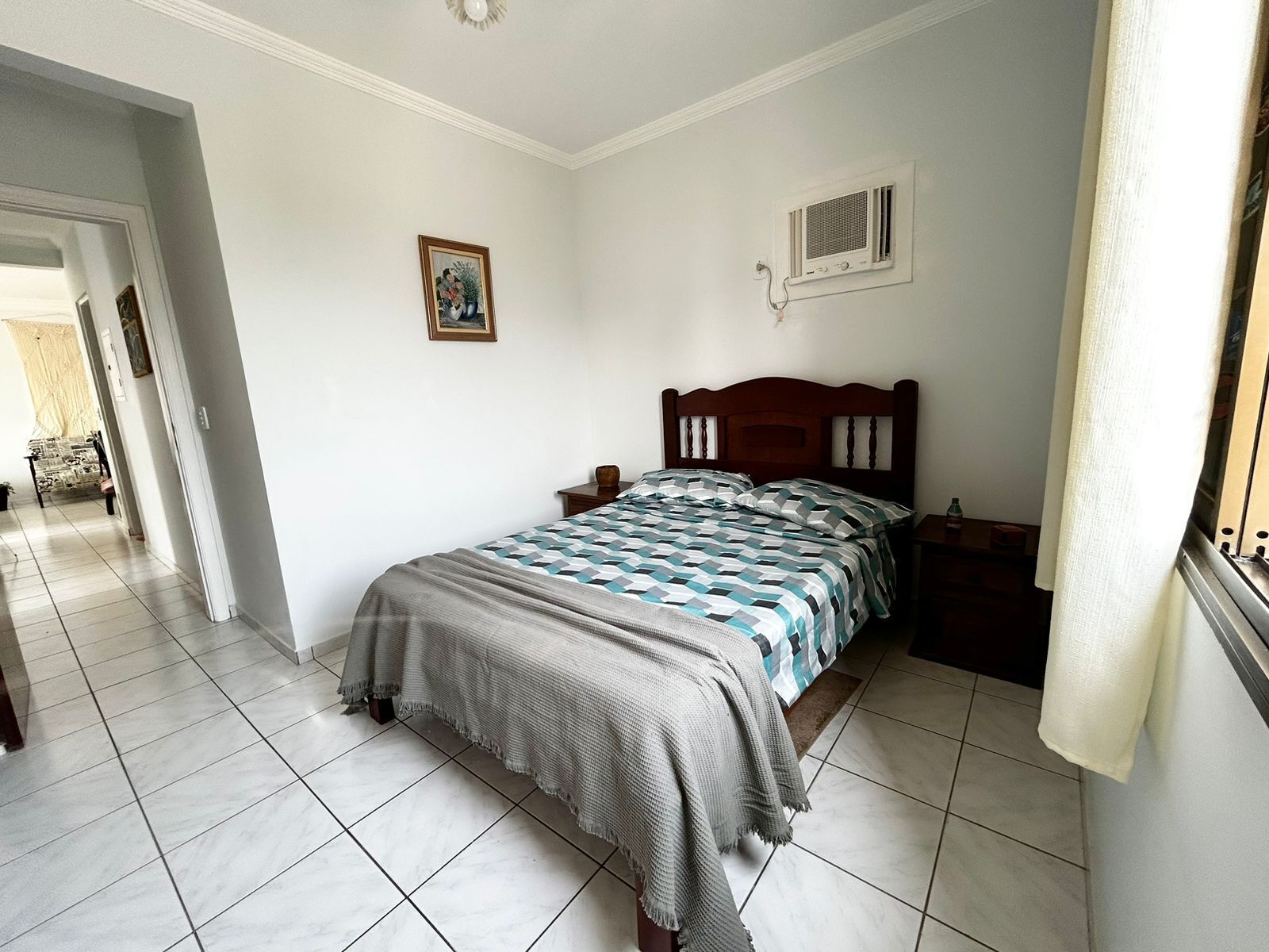 Quarto com cama, mesa de cabeceira, criadeira, um quadro na parede e janela com cortinas.