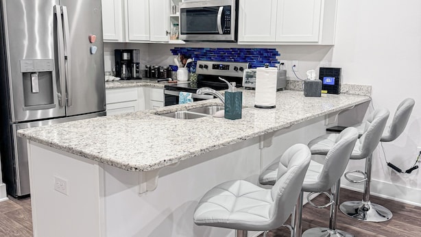 A modern kitchen with white cabinets, stainless steel appliances, a granite countertop, and a bar area with stools.