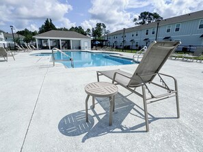 Zona de piscina con sillones de descanso y una pequeña mesa, rodeada por un edificio con un área de estar cubierta.
