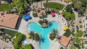 Vista aérea de um resort com uma piscina central, cercada por palmeiras, áreas de estar e um jardim bem cuidado.