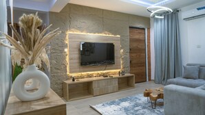 Ein modernes Wohnzimmer mit Steinwand, Flachbildfernseher, Holztische und einem grauen Sofa.