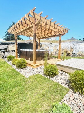 Een houten pergola met een geïntegreerd barbecuegedeelte, omringd door een aangelegde tuin met rotsen en struiken.