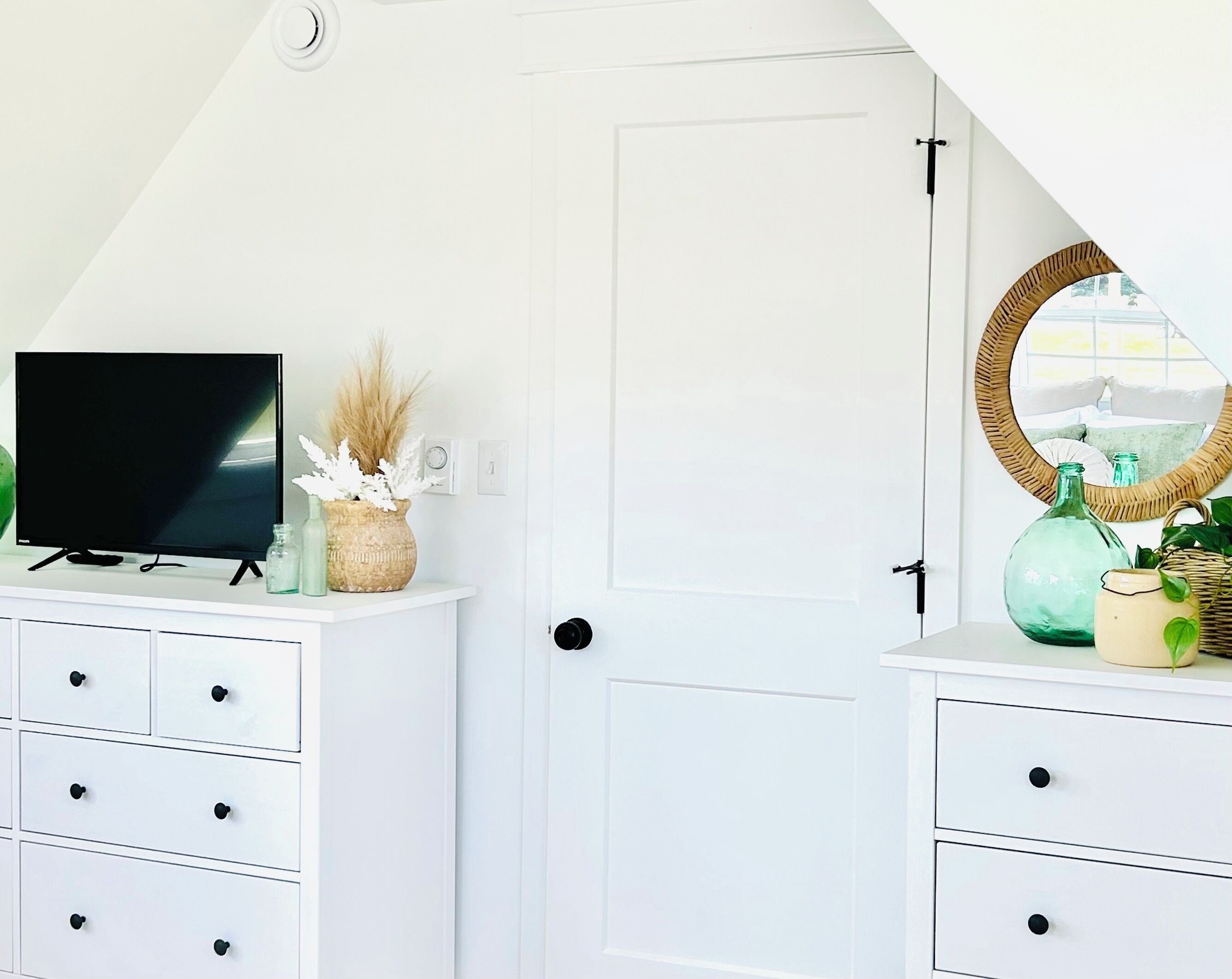 A room with a white door, a television on a stand, and a mirror with a wicker frame.