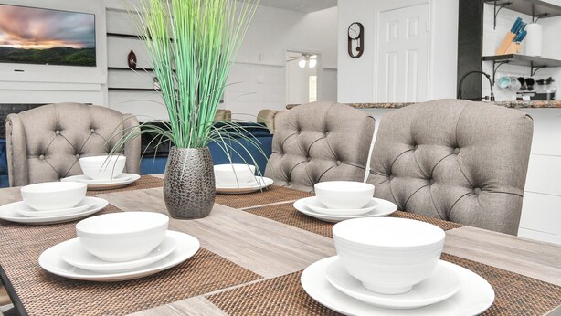 Un comedor con una mesa dispuesta para cuatro personas, rodeada de sillas capitonadas, una planta decorativa y un televisor al fondo.