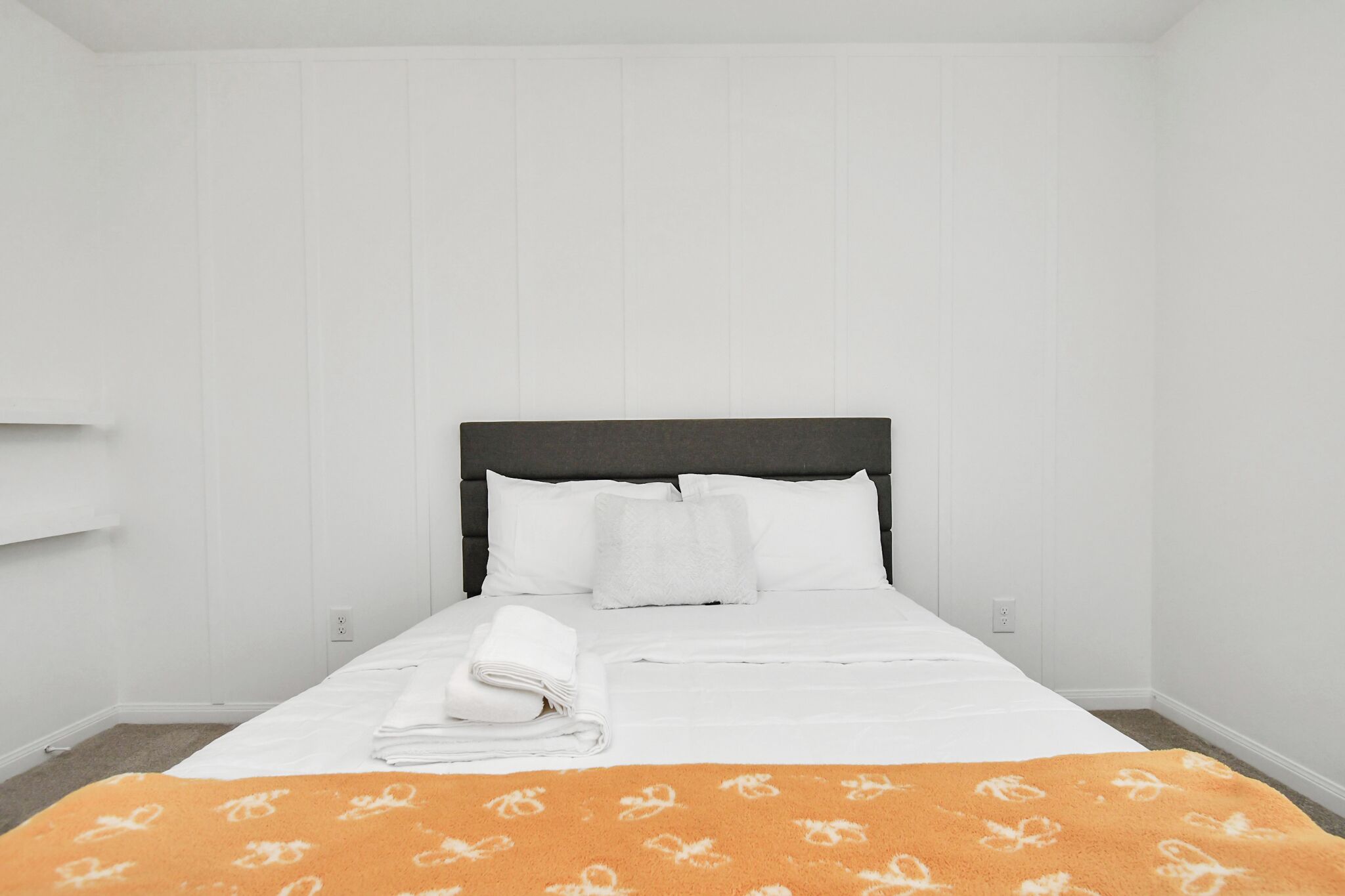 A neatly made bed with white linens and a folded white towel, a dark headboard, and a white wall with a light switch.