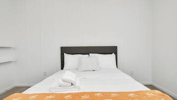 A neatly made bed with white linens and a folded white towel, a dark headboard, and a white wall with a light switch.