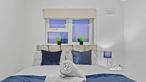 Une chambre à coucher bien rangée, avec un lit, deux fenêtres dotées de rideaux, une table de chevet avec une lampe et une petite plante en pot posée sur le rebord de la fenêtre.