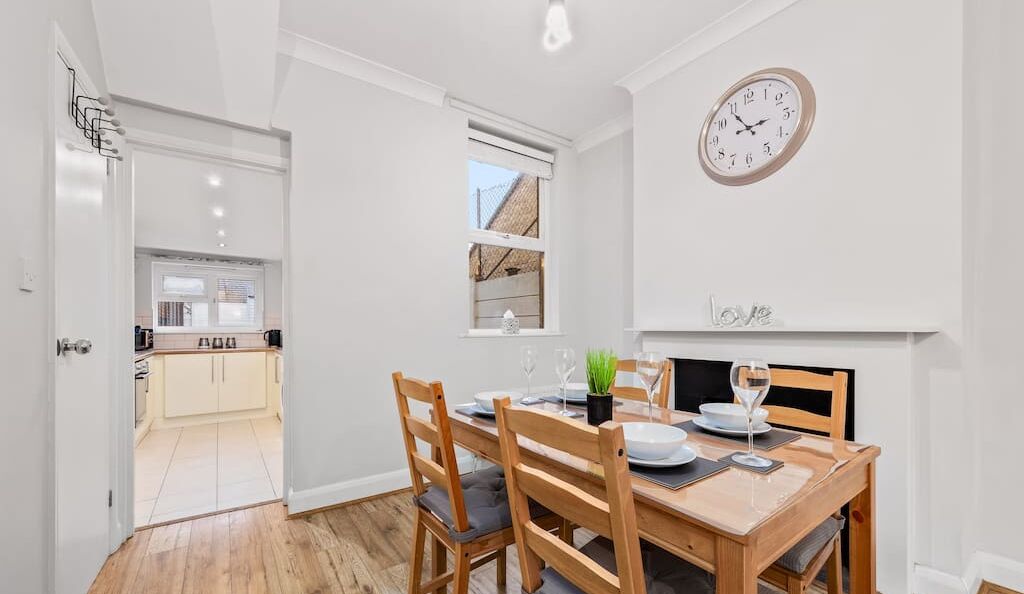 A dining area with a wooden table set for two, a clock on the wall, and a view into the kitchen.