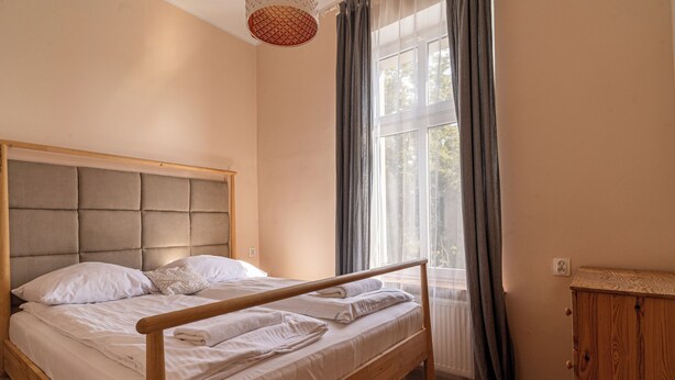 A neatly made bed with a wooden frame and headboard, a wooden nightstand, and a window with curtains. - Sand by Grand Apartments (Sopot)