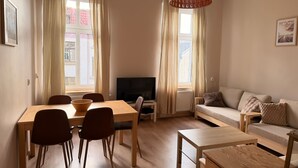 A living room with a dining table, chairs, a sofa, a TV, and a large window with curtains. - Sand by Grand Apartments (Sopot)