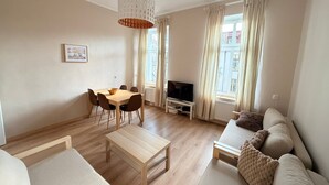 A modern living room with a wooden floor, a dining table with chairs, a sofa, a TV on a stand, and a hanging light fixture. - Sand by Grand Apartments (Sopot)