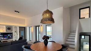 A modern living room with a round wooden dining table, grey sofas, and a hanging wicker chandelier. - Faragas Hill Oasis Villa (Aliki)