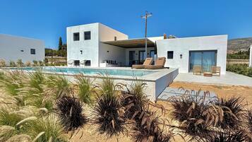 Une maison moderne à deux étages avec une piscine, entourée d’herbes sèches et d’arbustes.