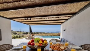 A breakfast table set with plates, cutlery, a fruit bowl, croissants, and juice glasses, overlooking a pool and landscape. - Faragas Hill Oasis Villa (Aliki)