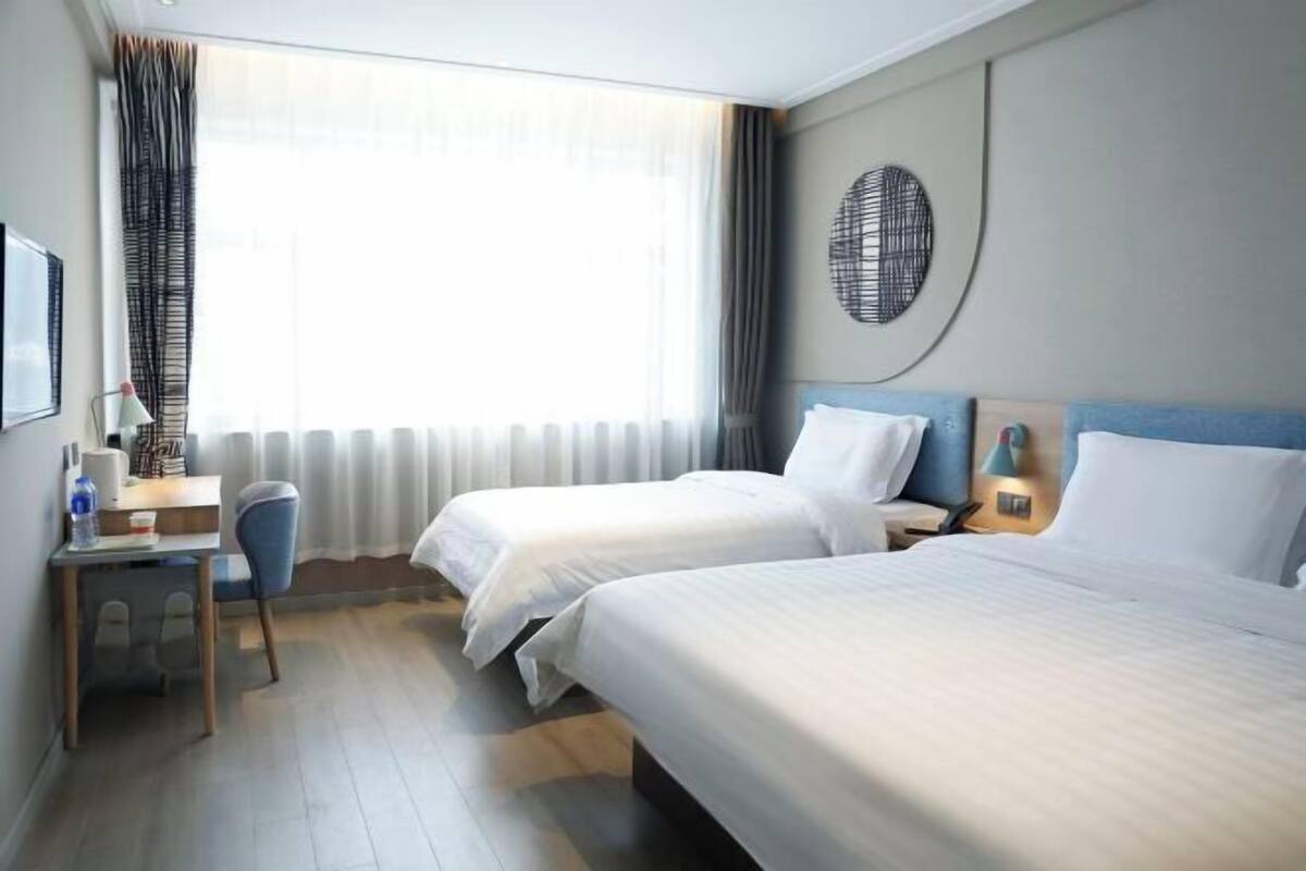Habitación de hotel con dos camas, un escritorio, una silla, una lámpara y un amplio ventanal con cortinas.