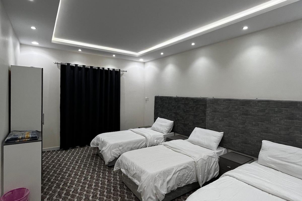 Ein modernes Hotelzimmer mit drei Einzelbetten, einem Kleiderschrank, einem Schreibtisch und einem gemusterten Teppich.